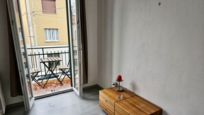 Et værelse med en trækommode, et lille bord med stole, og en balkon med udsigt til en gul bygning.