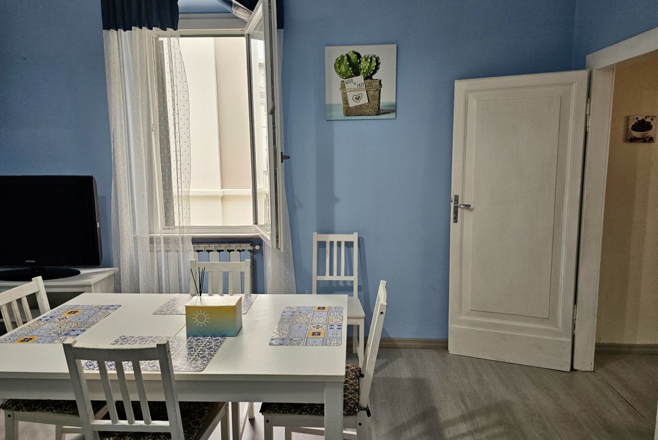Una habitación con una mesa de comedor, sillas, un televisor, una ventana con cortinas, una puerta y un cuadro colgado en la pared.