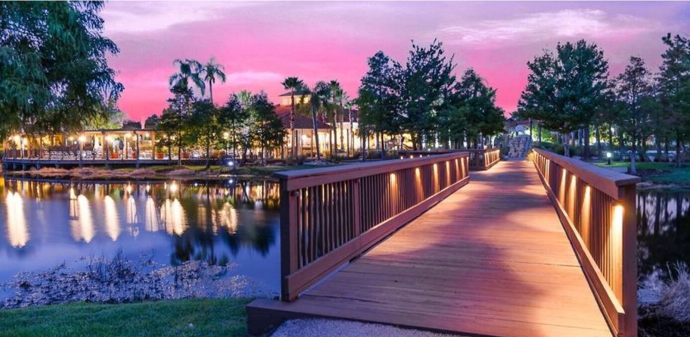 Un puente de madera sobre un cuerpo de agua tranquilo, con un cielo al atardecer, rodeado de árboles y un edificio iluminado.