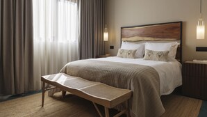 Quarto de hotel bem arrumado, com uma cama grande, uma mesa de cabeceira, uma poltrona para leitura e uma janela ampla com cortinas.