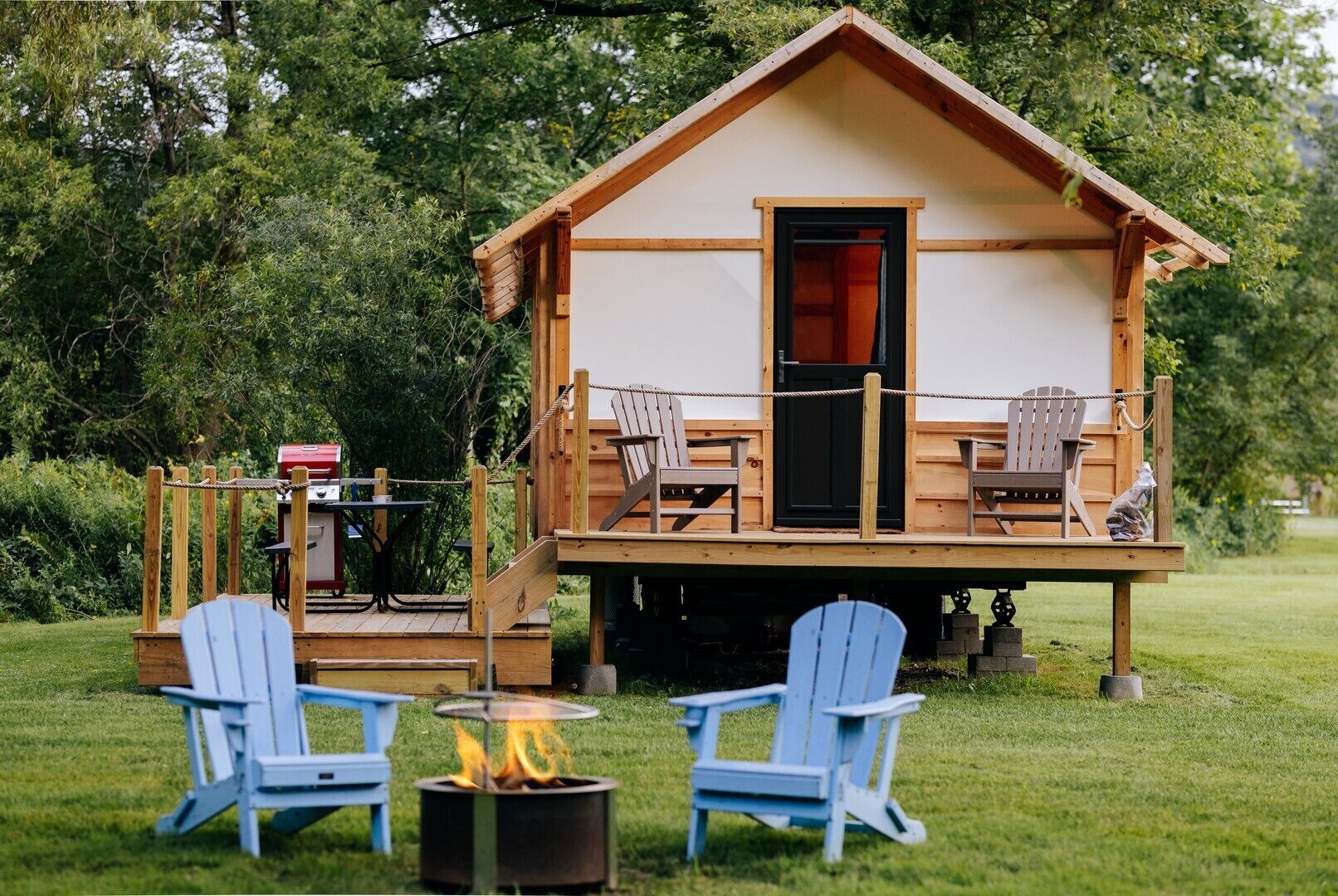 A wooden cabin with a deck, two Adirondack chairs, a fire pit, and a grill.