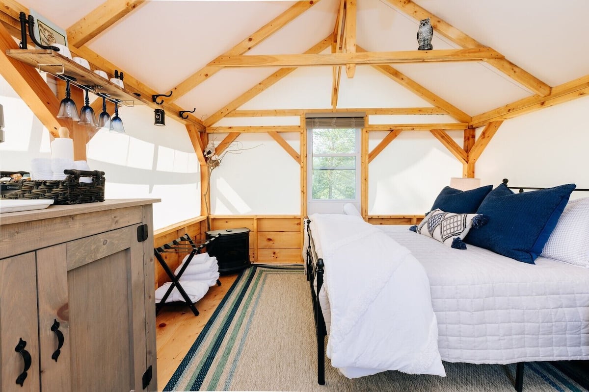 A cozy cabin bedroom with a bed, a wooden dresser, a window, and a rustic ceiling.