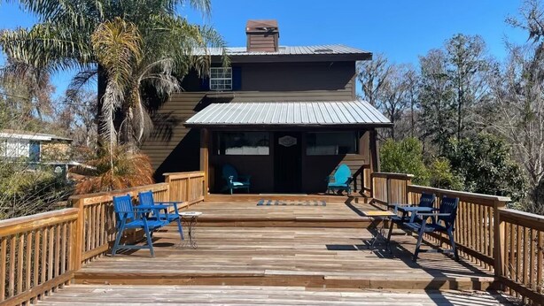 A wooden deck leading to a building with a metal roof, blue chairs, and a small table. - 3 Story Rainbow Riverfront House With Panoramic Views!  (Dunnellon)