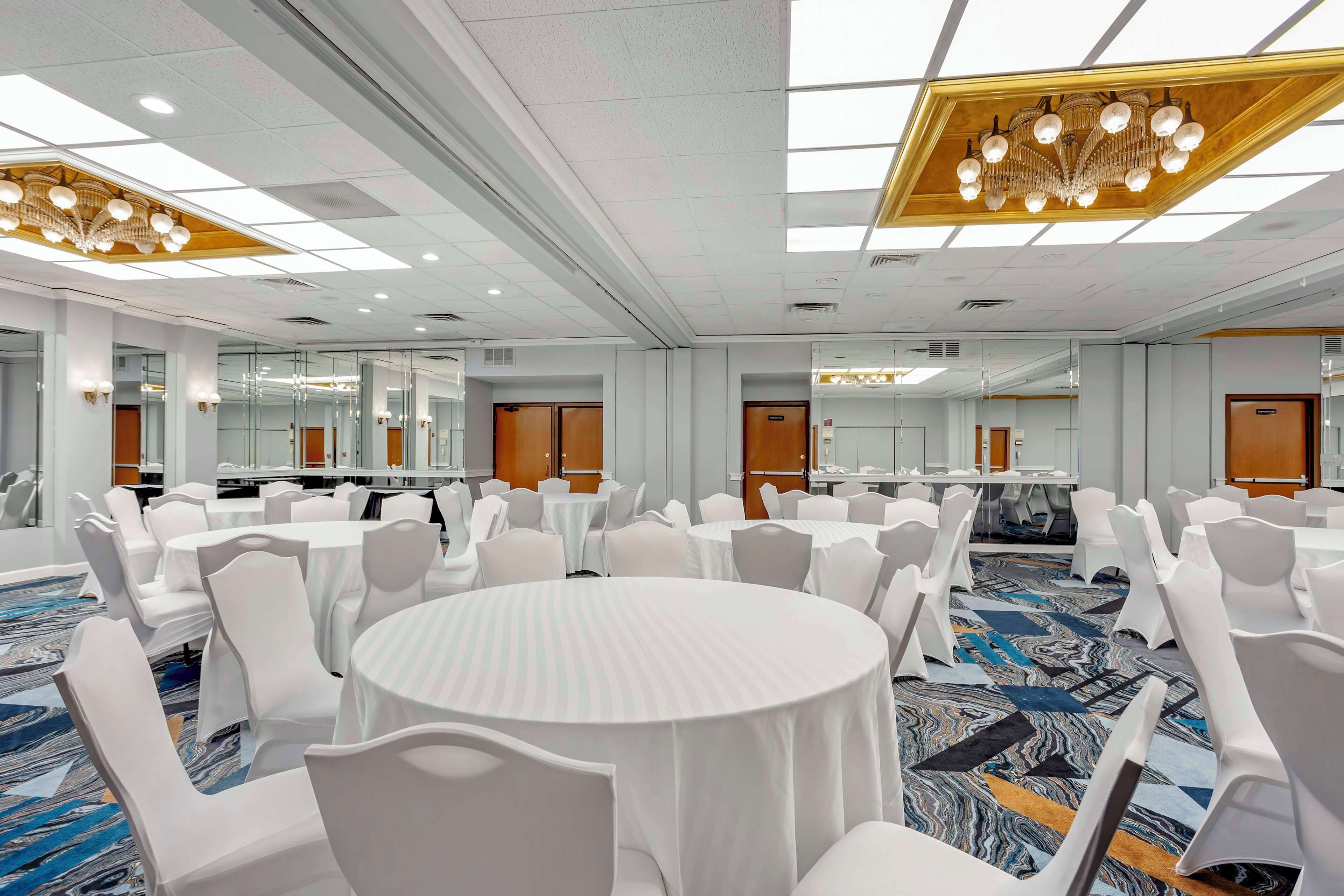 Une salle de conférence avec des tables rondes et des chaises blanches, de grandes fenêtres et un lustre.
