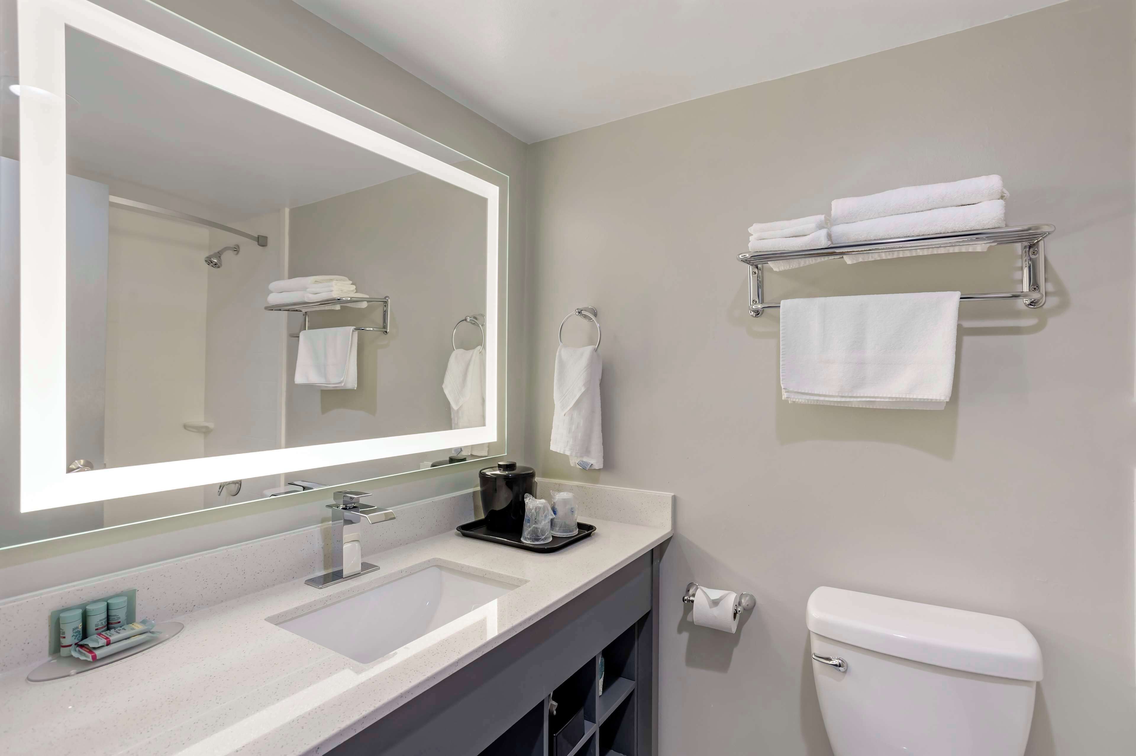 A modern bathroom with a large mirror, white sink, and a toilet. There are towels on a rack and a tray with toiletries.