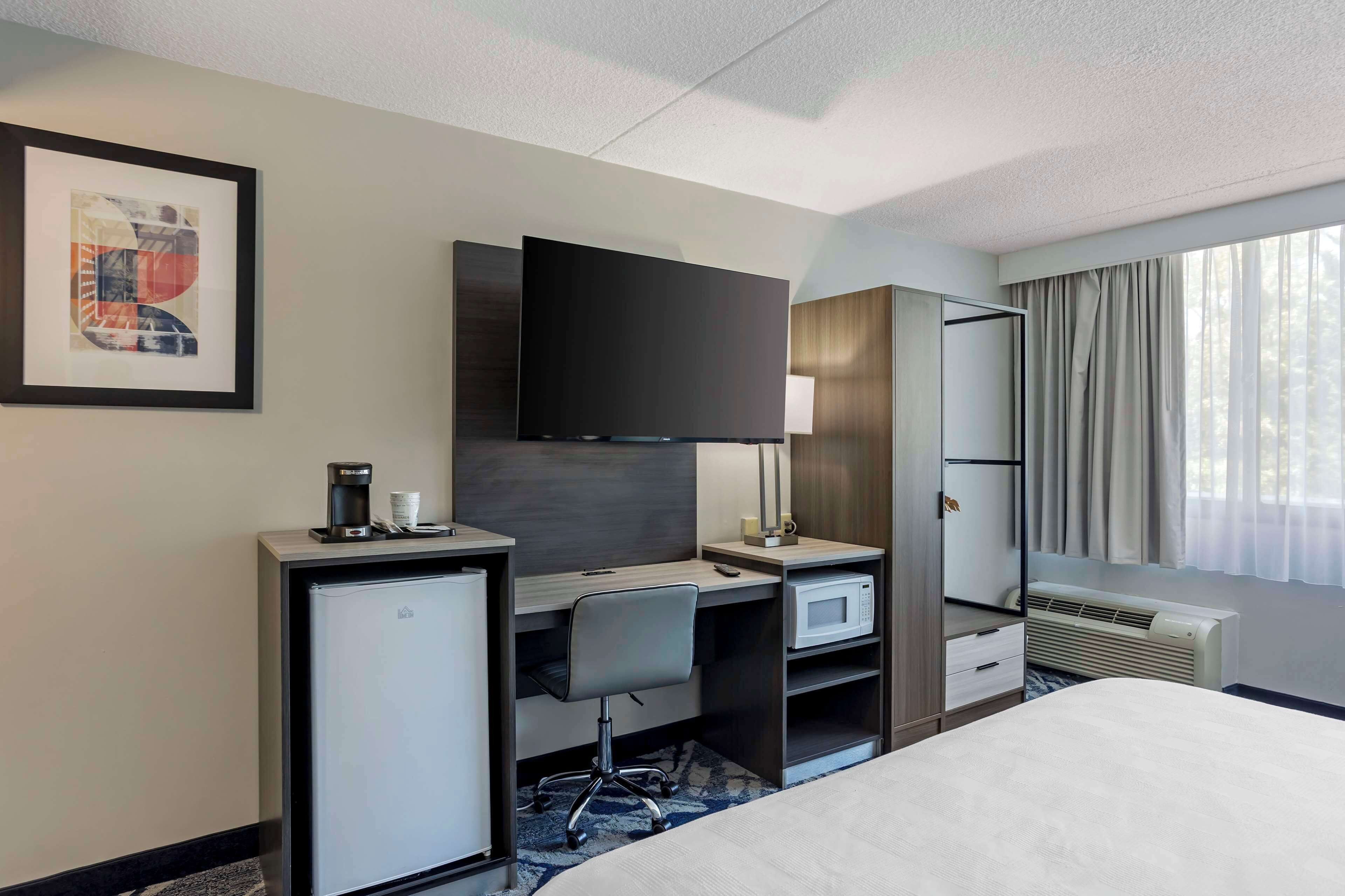 A hotel room with a bed, a desk with a chair, a flat-screen TV, a mini-fridge, a microwave, a heater, and a window with curtains.