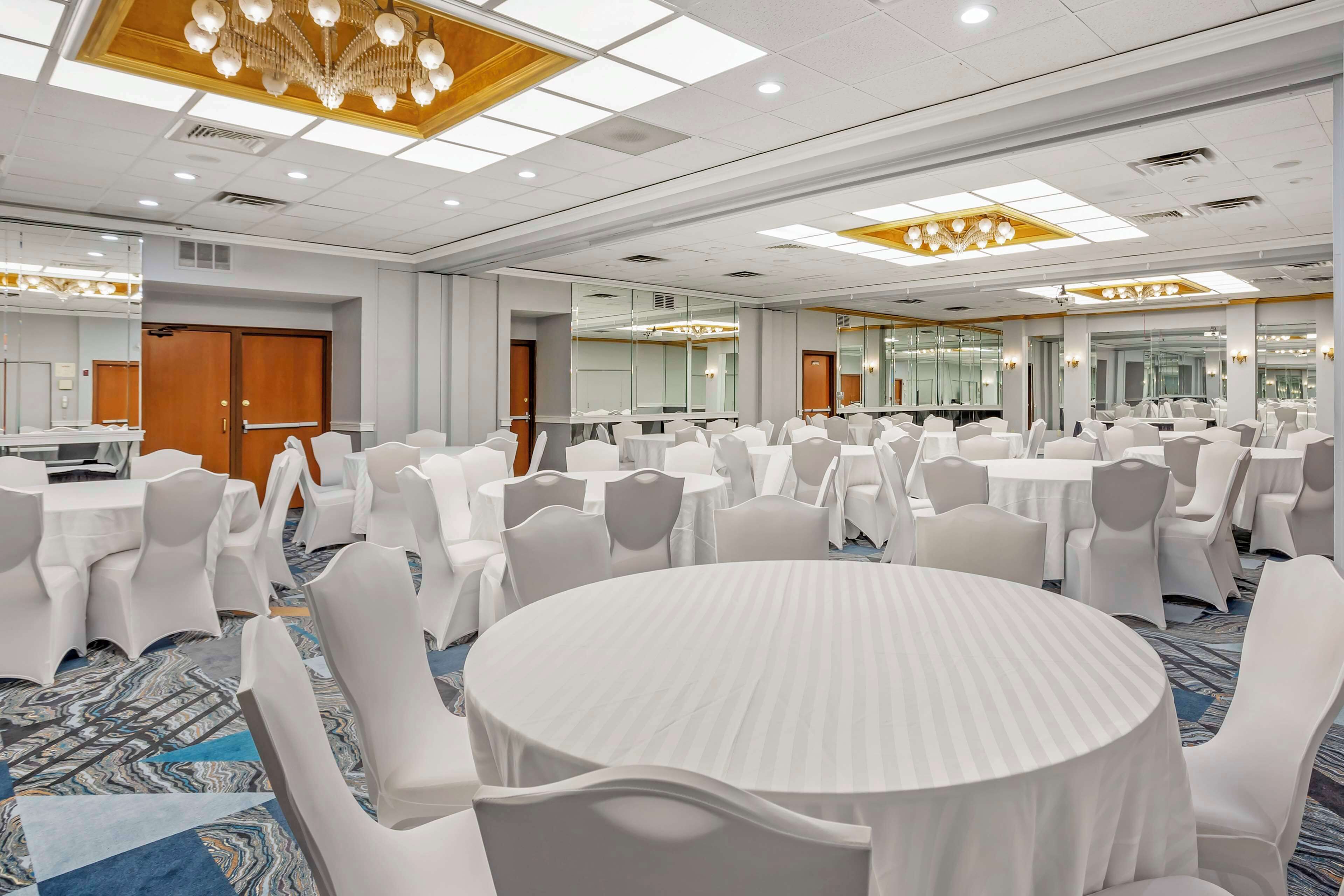 Une grande salle de conférence dotée de tables rondes et de chaises blanches, équipée de luminaires encastrés et d’un lustre.