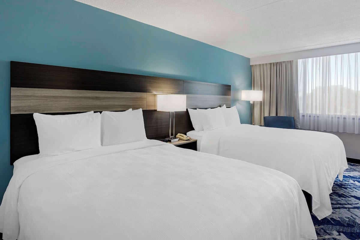 A hotel room with two beds, a nightstand, a lamp, and a view of the city through the window.
