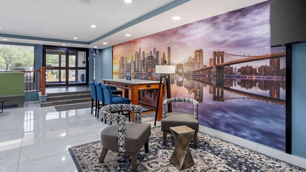 A hotel lobby with a large mural of a city skyline and a bridge, a reception desk, seating area with chairs, and a staircase leading to an outdoor area. - Best Western New Brunswick - East Brunswick (East Brunswick)