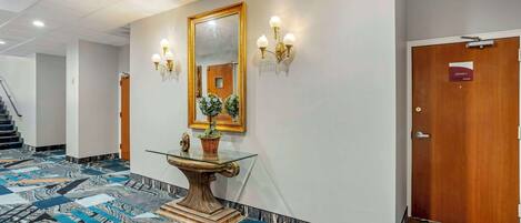 Un vestíbulo de hotel con piso de diseño de mármol, una mesa de vidrio con planta en maceta, un espejo grande, apliques de pared y una puerta de madera con la etiqueta "JERSEY".