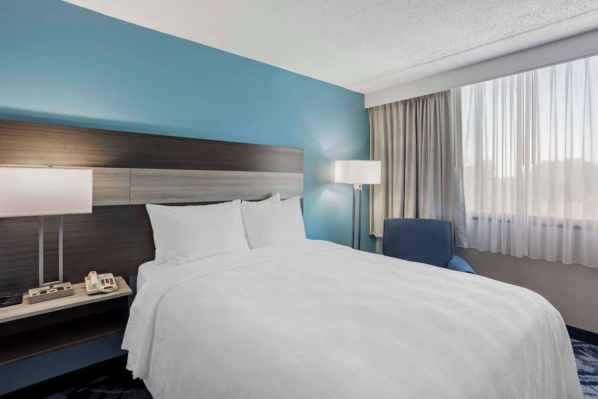 A hotel room with a large bed, a bedside table with a lamp, a phone, and a chair.