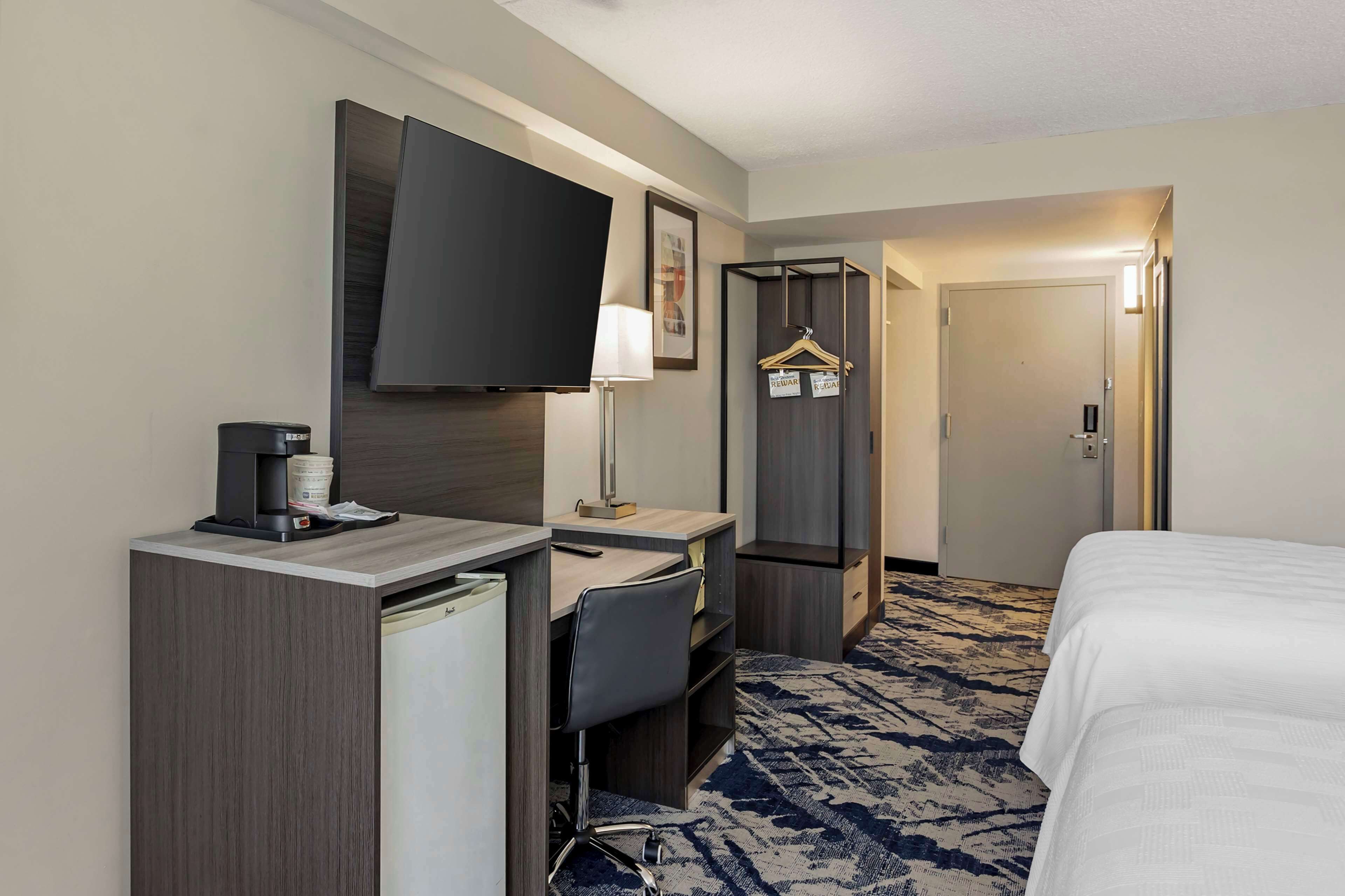 A hotel room with a bed, a desk with a chair, a television, a coffee maker, and a wardrobe.