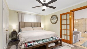 Ein Schlafzimmer mit Bett, Nachttischen, Deckenventilator, einer Tür mit Milchglasfenstern und Blick ins Bad.