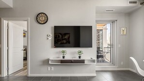 A modern living room with a flat-screen TV mounted on the wall, a white console table with two potted plants, a large clock, and a glass coffee table. - City View 1BR Downtown Calgary (Calgary)