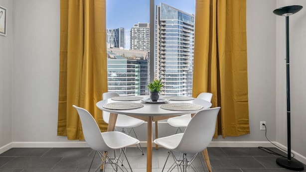 A modern dining area with a round table, white chairs, and a view of city buildings through large windows with yellow curtains. - City View 1BR Downtown Calgary (Calgary)