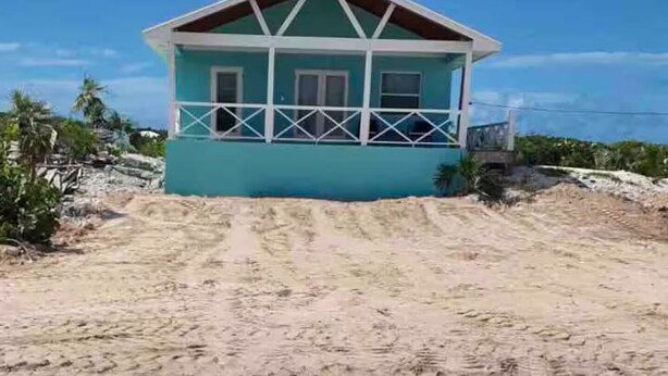 Une maison de plain-pied avec un toit à pignon, un balcon et une allée menant à la maison recouverte de sable.