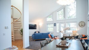 A spacious living area with a wooden staircase, a blue sofa, a dining table with chairs, and large windows providing natural light.