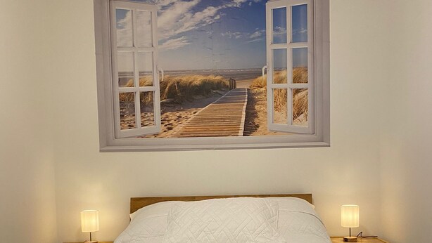 Ein Bett mit Holzrahmen, zwei Nachttische mit Lampen und ein an der Wand hängendes Bild mit einer Strandansicht.