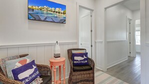 A hallway with a television displaying a beach scene, wicker chairs with 'BEACH HOUSE' pillows, and a small table with a decorative item. - Mollie Sunshine 117 SEV (Port Aransas)