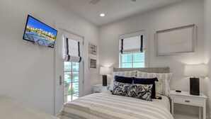 A bedroom with a bed, two bedside tables, a TV mounted on the wall, a window with blinds, and a door with a view of a pool. - Mollie Sunshine 117 SEV (Port Aransas)