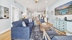 A spacious living room with a blue sofa, a ceiling fan, a large painting, and a dining area with a table and chairs. - Beach Haven TC 3103 (Port Aransas)