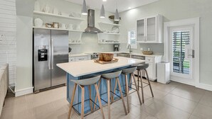 A modern kitchen with a stainless steel refrigerator, white cabinets, a central island with stools, and pendant lighting. - Cloud Nine 136 BE (Port Aransas)