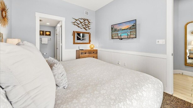 Ein Schlafzimmer mit einem Bett, einer Kommode, einem Spiegel, einem an der Wand hängenden Bild und einer Tür zu einem anderen Raum.