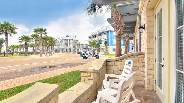A hotel with a stone wall patio, outdoor seating, and a view of a street with palm trees and a golf cart. - Suntide TC 1103 (Port Aransas)