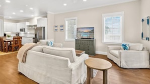 A spacious living area with a sofa, a coffee table, and a TV. There is a kitchen in the background with white cabinets and a refrigerator. - Palm Tree Retreat TC 3102 (Port Aransas)