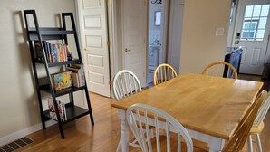 A dining area with a wooden table and white chairs, a black bookshelf with books, and a white door leading to a bathroom.