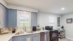 Una cocina compacta con armarios grises, un lavavajillas de acero inoxidable, una estufa negra y una ventana con persianas.