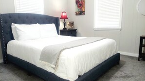Una cama con ropa blanca, cabecera oscura, lámpara roja y una pequeña señal de madera en una mesita de noche.