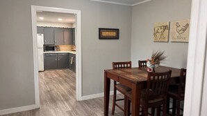 Un comedor con una mesa y sillas de madera, una cocina al fondo y un cuadro en la pared.