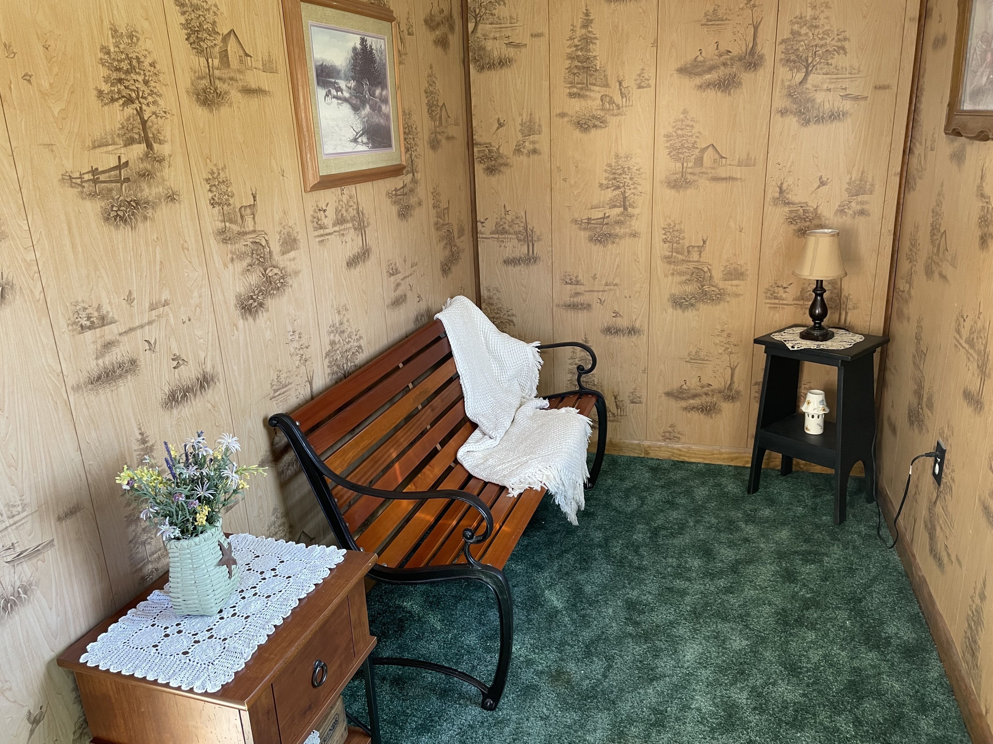 Una habitación con un banco de madera, una mesita con un florero, un cuadro en la pared y una mesita de noche con una lámpara.