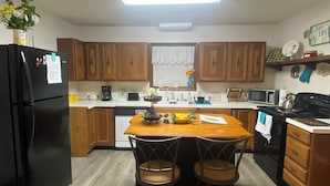 A kitchen with wooden cabinets, a black refrigerator, a wooden dining table with two chairs, a microwave, a stove, and a window with a curtain.