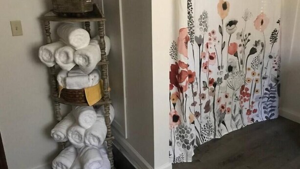 A room with a wooden shelf holding rolled towels and a floral curtain.
