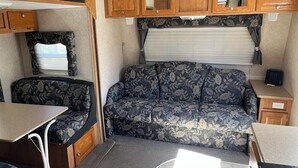Interior de una caravana compacta con sofá, una mesita, armarios de madera y una ventana con cortinas.