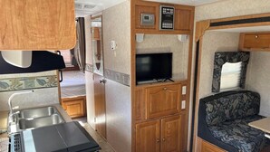 Interior de una caravana compacta con kitchenette, un televisor de pantalla plana, una zona de estar con un cojín estampado y un mueble pequeño con un reloj.