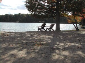 A lakeside view with two Adirondack chairs under a tree, a sandy area, and a calm lake. - The Rockwood Trailer/RV - Sandy Beach + Free Watercraft Use in a Natural Setting (Cloyne)