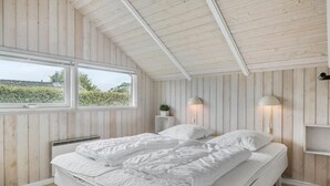 Ett sovrum med ett sluttande tak, träpanel på väggarna, ett stort fönster, två sänglampor och en säng med vita sängkläder.