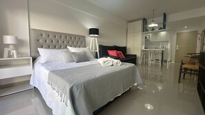 Quarto moderno com cama grande, mesas de cabeceira, escrivaninha e pequena cozinha.