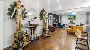 A spacious living area with a wooden floor, a dining table, and a sofa. There are two decorative statues on pedestals. - Spacious open floor plan. 5 beds. 5 TVs. & Art. (Fayetteville)