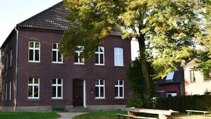 A two-story brick building with a central entrance, a small garden area with a bench, and a tree in front. - Ferienwohnung Josef - Wohnen wie zur Gründerzeit (Kevelaer)