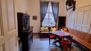 A room with a wooden floor, a dining table with chairs, a sofa with cushions, a television on a cabinet, and a window with curtains. - Ferienwohnung Josef - Wohnen wie zur Gründerzeit (Kevelaer)