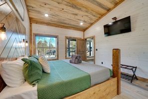 Un dormitorio tipo cabaña de madera con una cama, ropa de cama verde y blanca, un televisor montado en la pared y ventanas amplias.