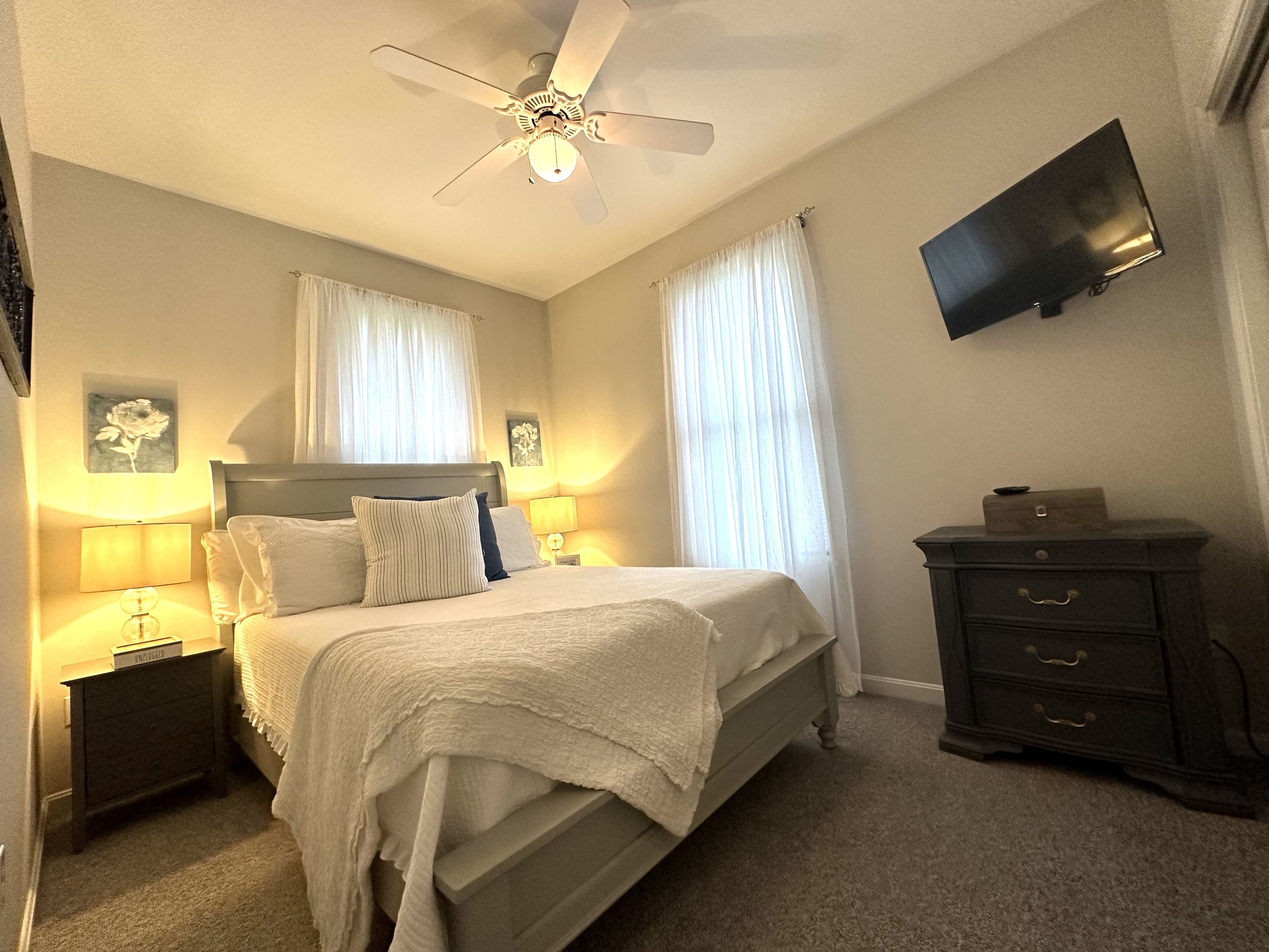 Un dormitorio con cama, mesitas de noche, cómoda, ventilador de techo y televisor de pantalla plana.