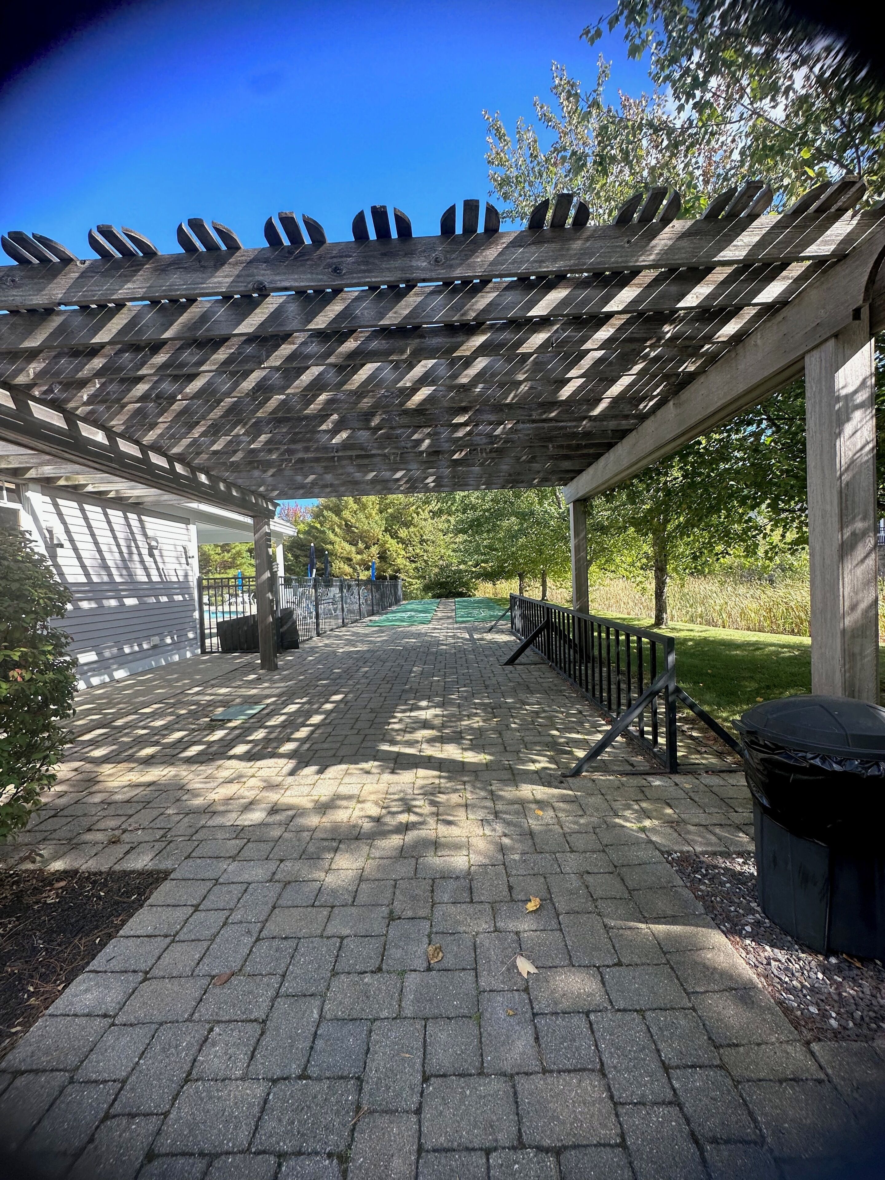 A covered outdoor area with a brick pathway, a pergola with slats, and a trash bin.