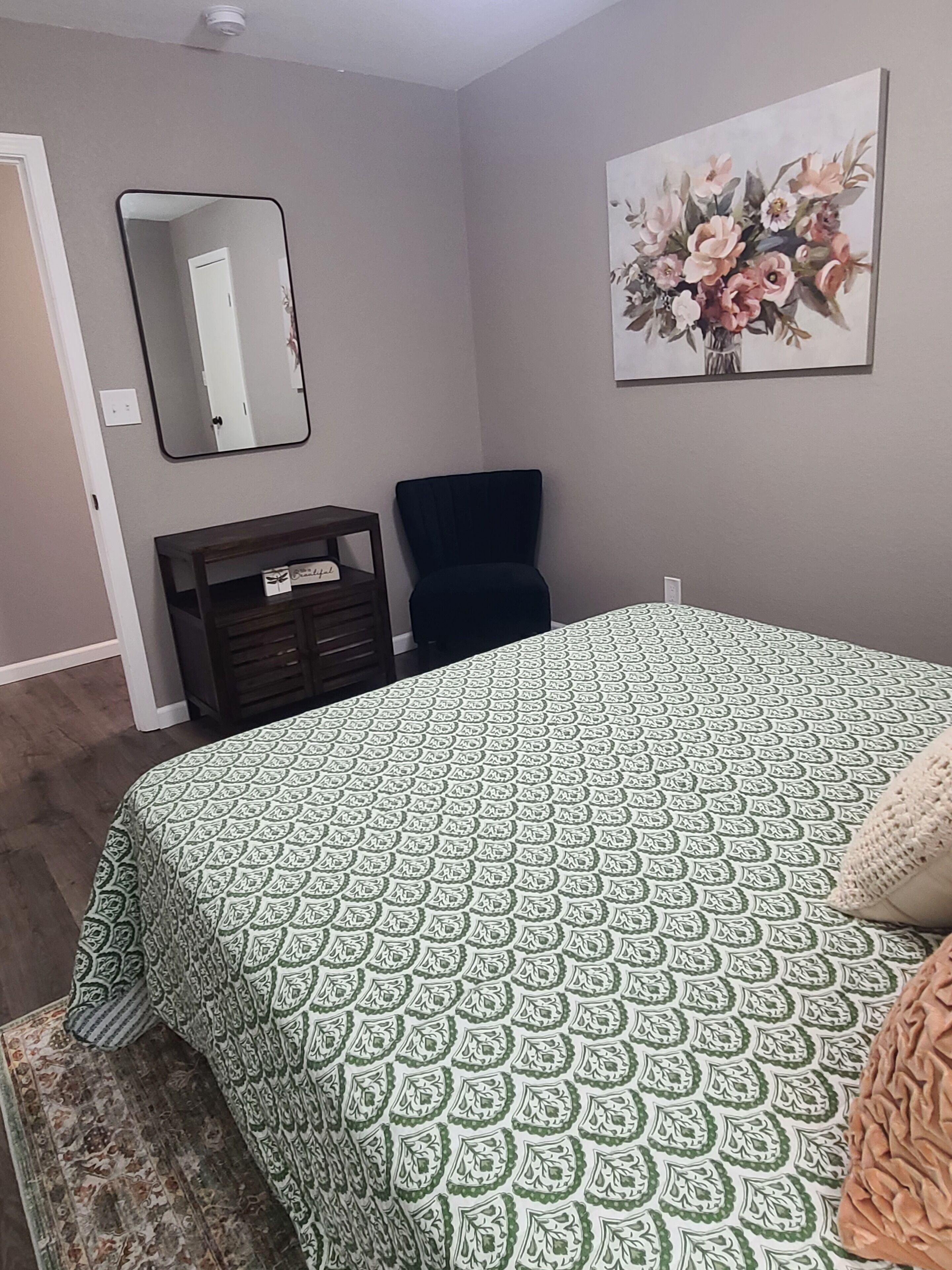 A bedroom with a patterned bedspread, a dark armchair, a dresser, a mirror, and a floral painting on the wall.