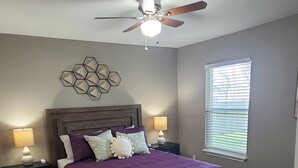 A bedroom with a bed, two bedside tables, a ceiling fan, a window with blinds, and a decorative wall piece. - Pet-Friendly Home for 7 with Fire Pit (Junction)