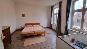 Une chambre à coucher avec un lit en bois, une table de chevet, une commode, une fenêtre avec des rideaux et une vue sur les bâtiments à l’extérieur.